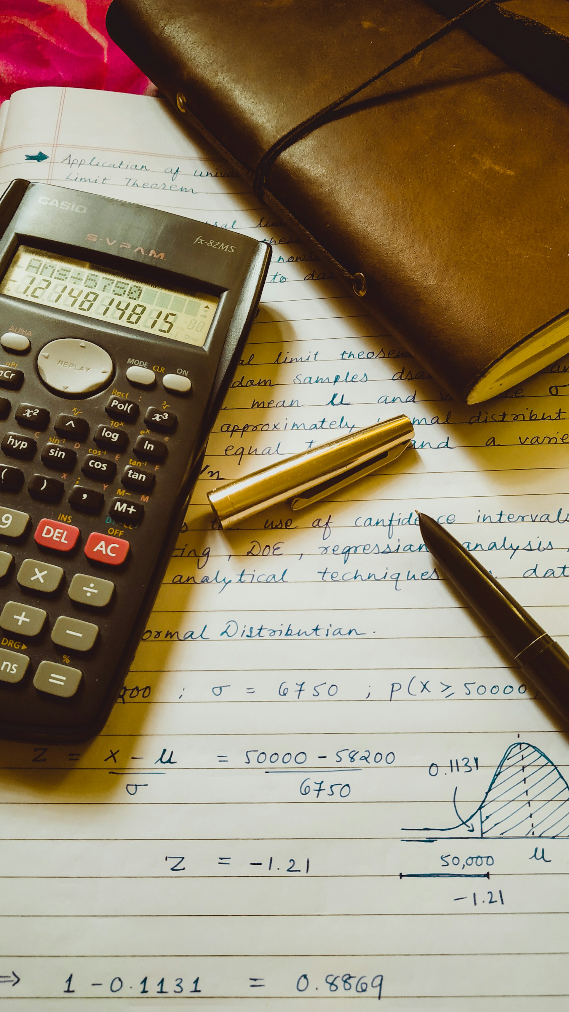 A scientific calculator, handwritten technical notes with statistical calculations, and a leather notebook on a desk
